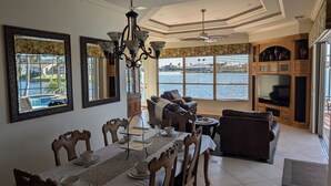 A dining area with a view of a body of water, a chandelier, and a television in a wooden cabinet. - CasaSuLago Sunset Lakefront, Saltwater Pool & Spa, Fishing, Kayaks & Game Room! (Cape Coral)