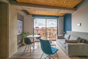 Un salon moderne avec un canapé, une table à manger et des chaises. Une grande fenêtre offre une vue sur les immeubles.