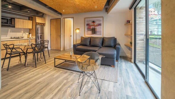A modern living room with a sofa, coffee table, and chairs. There is a kitchen area with a refrigerator and microwave. The room has large windows and a wooden ceiling. - NOK Stunning 1 Br in Chapinero (Bogotá)