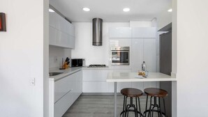 A modern kitchen with white cabinetry, a stainless steel range hood, a built-in oven, and two bar stools.