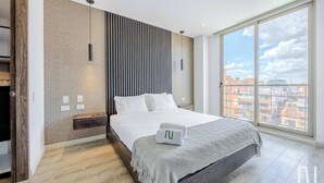 A modern bedroom with a large bed, a wooden bench, a wall-mounted light, and a view of buildings through a large window.