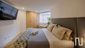A modern hotel room with a large bed, a TV mounted on the wall, a bench, and a window with a view of the city. - NOK Stylish 3BR Apt & Fast Wi-Fi Bogotá (Bogotá)