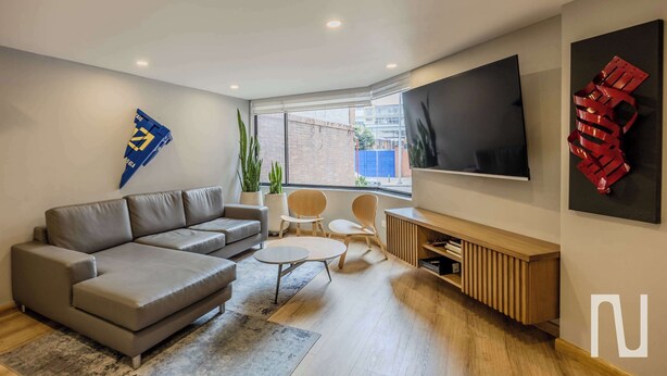 A modern living room with a grey sofa, a coffee table, and a TV mounted on the wall. There are two chairs and a small table. - NOK Stylish 3BR Apt & Fast Wi-Fi Bogotá (Bogotá)