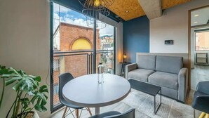 Ett modernt rum med ett runt bord, en soffa, en krukväxt och utsikt över en byggnad med tegelstensfasad genom stora fönster.