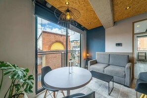 Une pièce moderne avec une table ronde, un canapé, une plante en pot et une vue sur un bâtiment en briques, visible à travers de grandes fenêtres.