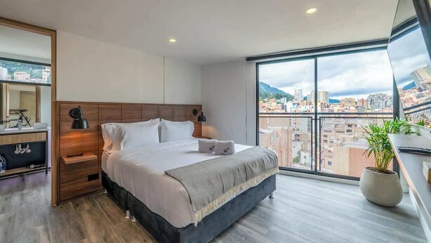 Un dormitorio moderno con una cama grande, mesitas de noche, un espejo y vistas a la ciudad a través de amplias ventanas.