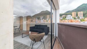 Una sala de estar moderna con sofá, mesa de centro y sillas, ventanas amplias con vista a la ciudad y un balcón con vistas a edificios y zonas verdes.