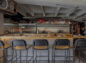 Un espace bar doté d’un comptoir en acier inoxydable, de plusieurs tabourets de bar et d’une variété de bouteilles et de verres disposés sur des étagères.