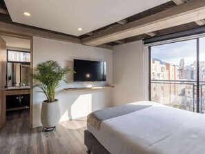 Een moderne slaapkamer met een groot bed, een aan de muur bevestigde televisie, een potplant en uitzicht op de stad.