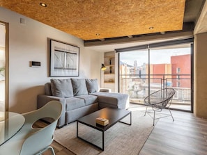 Een moderne woonkamer met een grijze hoekbank, een glazen salontafel, een metalen stoel en een grote schuifpui naar een buitenbalkon.
