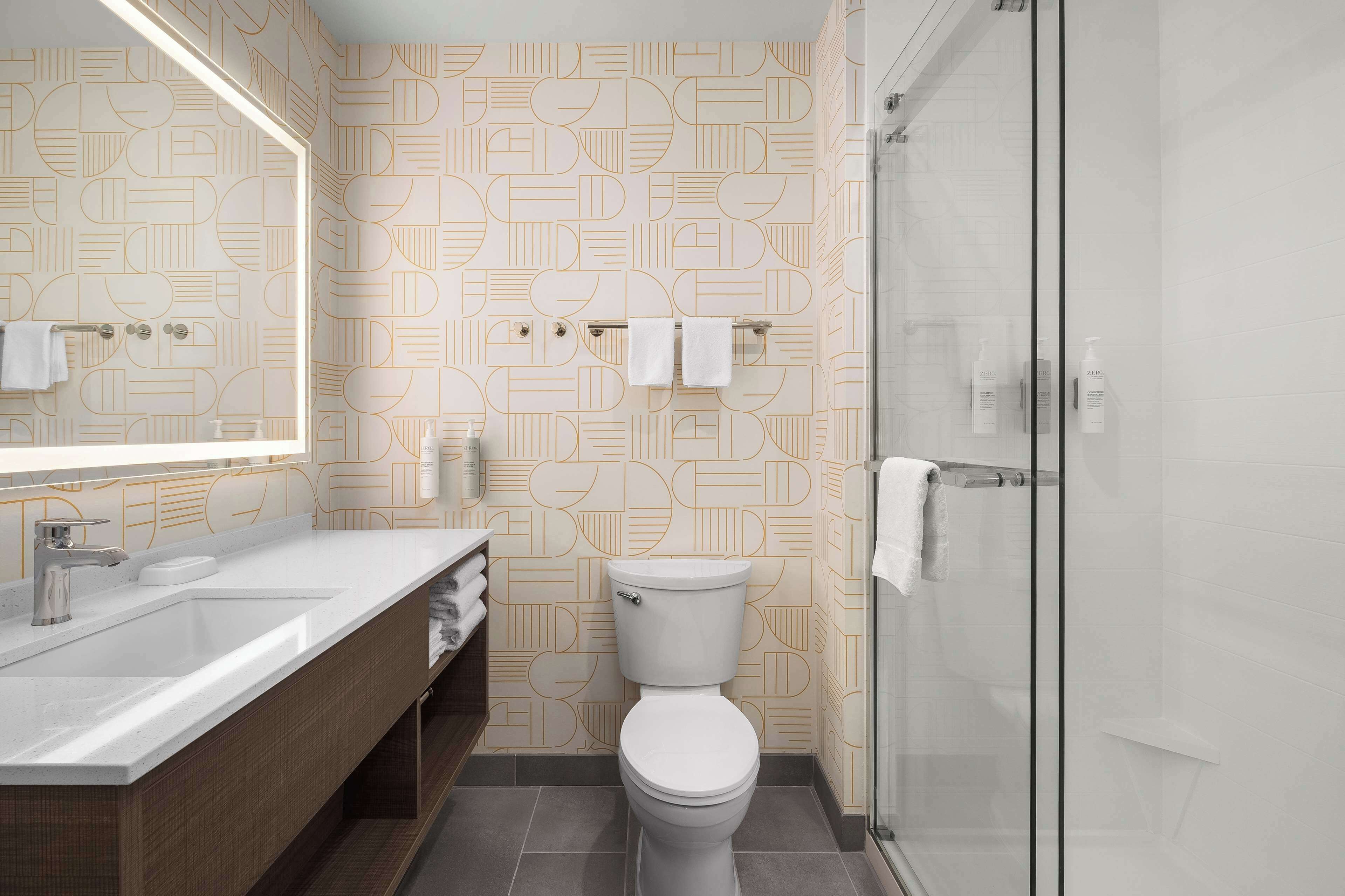 Une salle de bain moderne avec un mur orné d’un motif géométrique, un lavabo blanc, des toilettes et une douche à parois de verre.