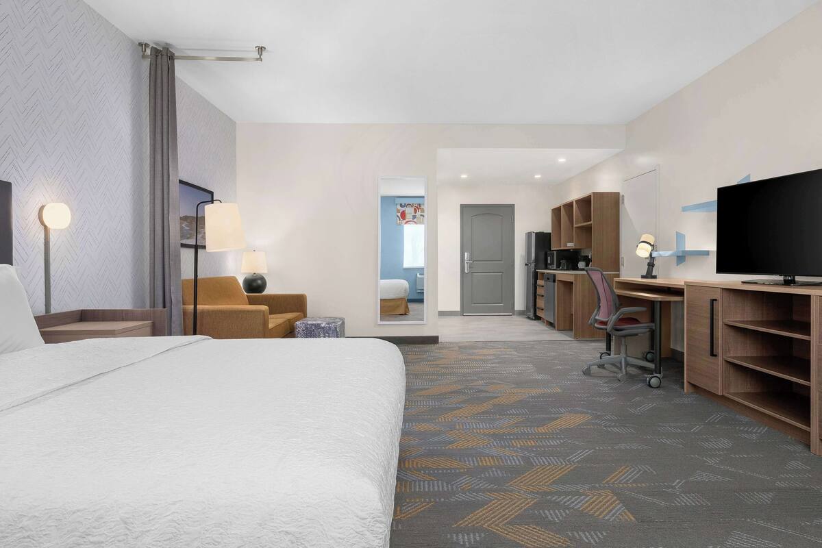 Ein Hotelzimmer mit einem großen Bett, einem Schreibtisch mit Computer, einem Fernseher und einem Sessel.