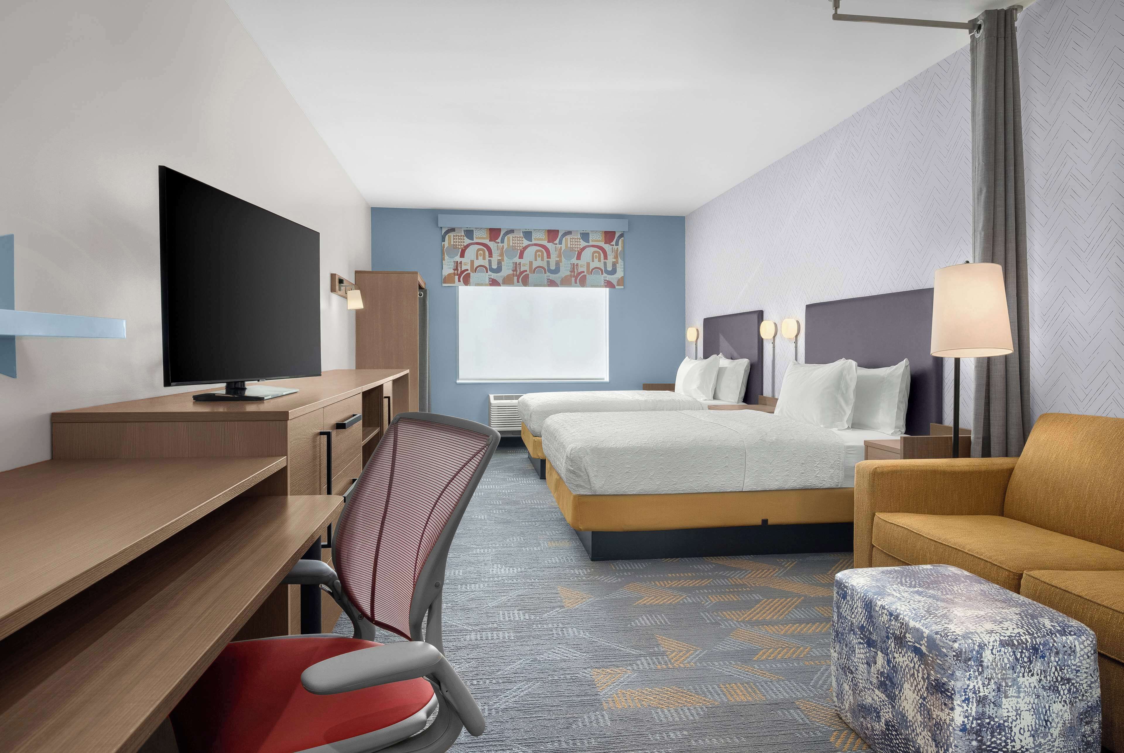 A hotel room with a desk, two beds, a TV, a chair, and a lamp.