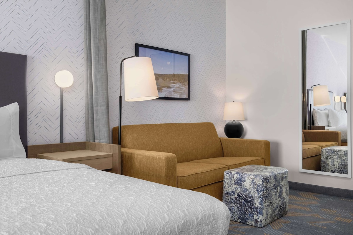 Ein Hotelzimmer mit einem großen Bett, einer Couch, einem kleinen Hocker, einem Nachttisch, einer Lampe, einem gerahmten Bild und einem Spiegel.
