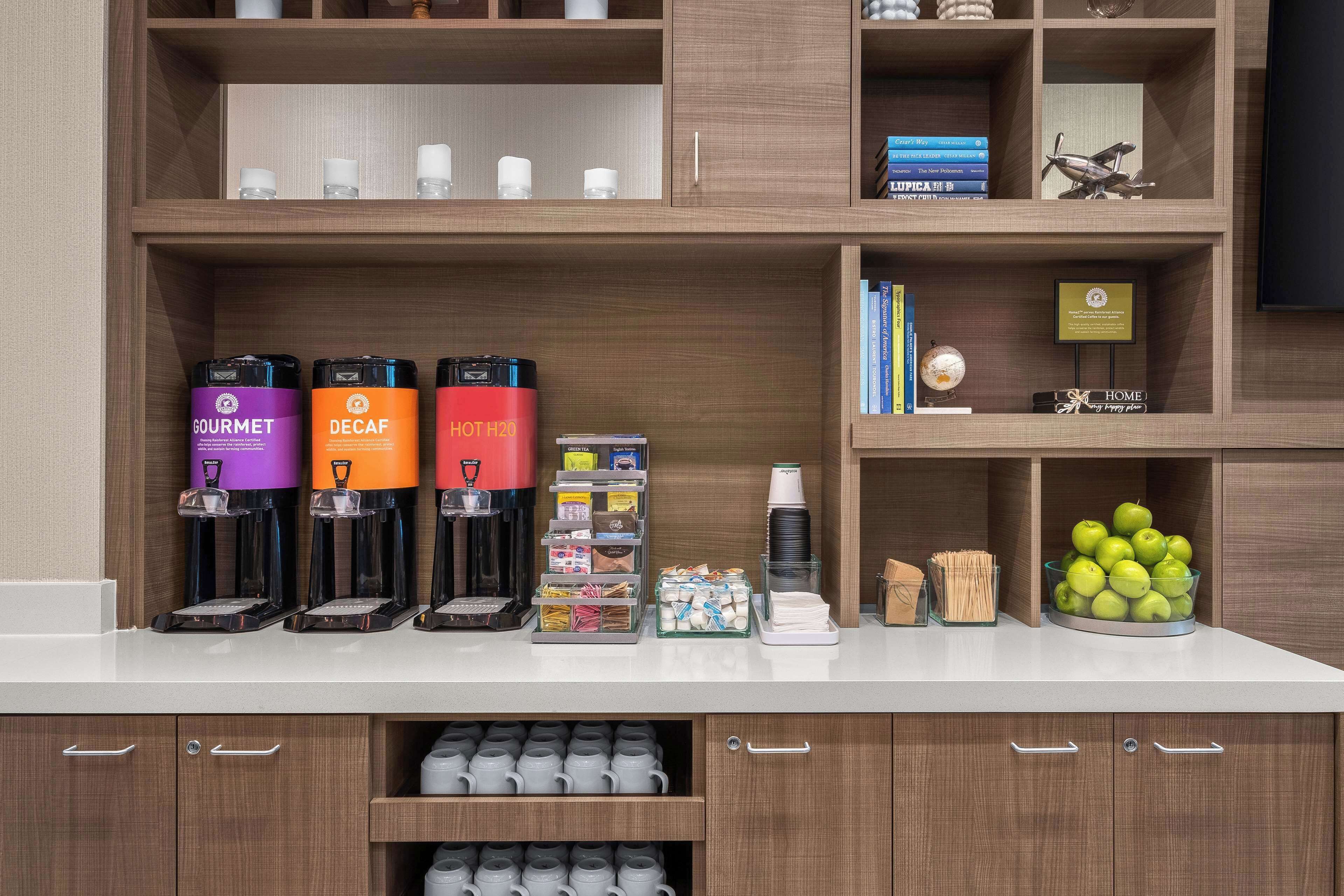 Un coin café équipé de trois machines à café, étiquetées respectivement « GOURMET », « DÉCAFÉINÉ » et « EAU CHAUDE ». On y trouve également divers dosettes de café, une petite pile de tasses et un bol de pommes sur le comptoir.