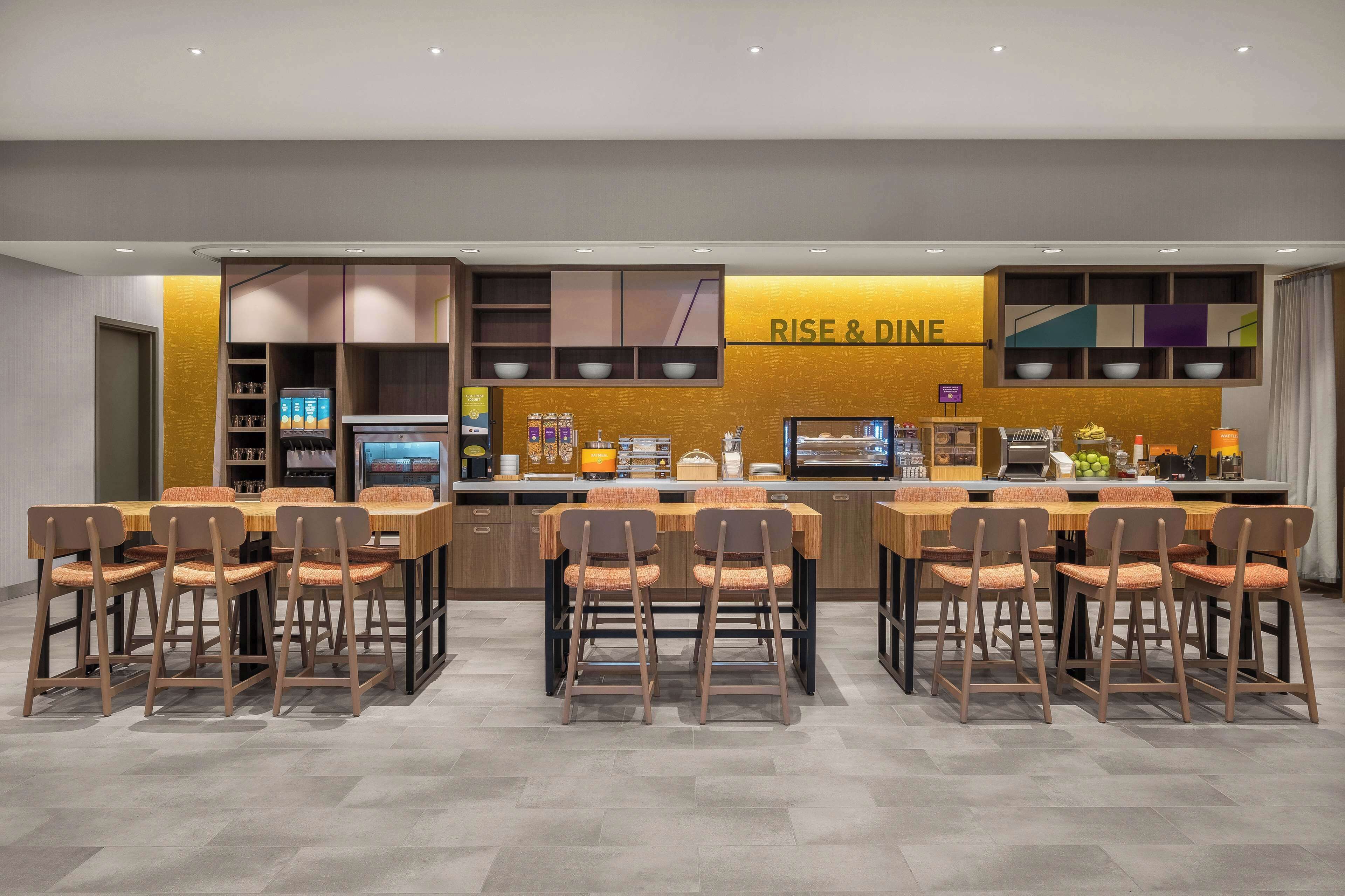 Un comedor moderno con una mesa larga y múltiples sillas, una encimera de cocina con un cartel que dice 'RISE & DINE', y una variedad de electrodomésticos y utensilios de cocina.