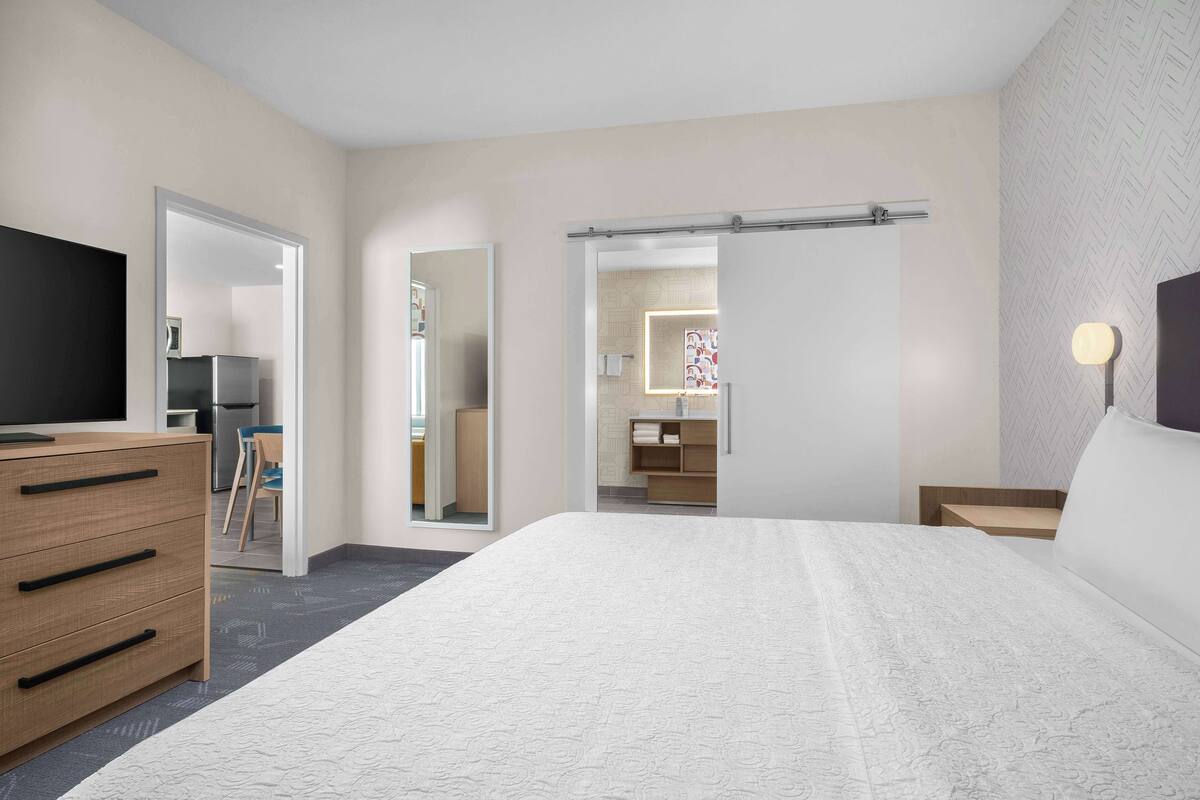 Ein modernes Hotelzimmer mit einem großen Bett, einer Holzkommode, einem Fernseher und einem durch eine offene Tür einsehbaren Badezimmer.