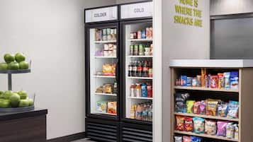 Una sección de aperitivos y bebidas con dos refrigeradores, uno etiquetado 'COLDER' y otro 'COLD', y una estantería llena de diversos snacks y bebidas.