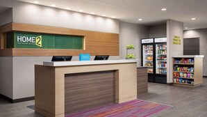 A reception desk with a sign reading "HOME 2 Suites by Hilton," a minibar, a refrigerator labeled "COLDER," and a snack shelf. - Home2 Suites By Hilton Winter Haven (Winter Haven)