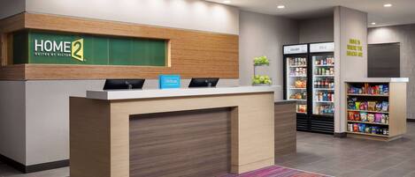 Un comptoir d’accueil avec une enseigne indiquant « HOME 2 Suites by Hilton », un minibar, un réfrigérateur portant l’inscription « PLUS FROID » et une étagère à collations.