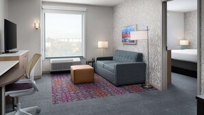 A hotel room with a desk, chair, sofa, ottoman, and a large window. - Home2 Suites By Hilton Winter Haven (Winter Haven)