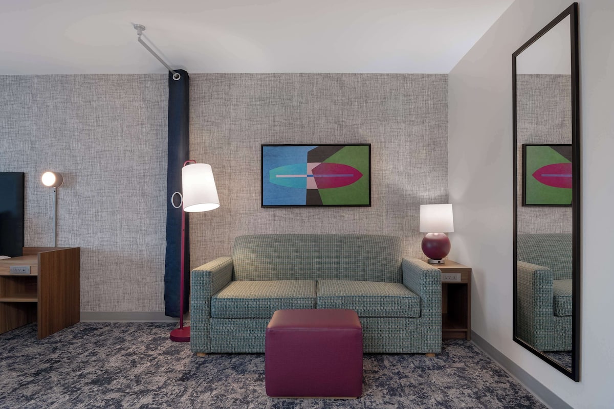 Een hotelkamer met een groene bank, een rode poef, een grote spiegel en een tv aan de muur.