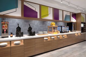 Una barra de desayuno moderna con diversos alimentos y electrodomésticos, incluyendo una cafetera y una exhibición de frutas.
