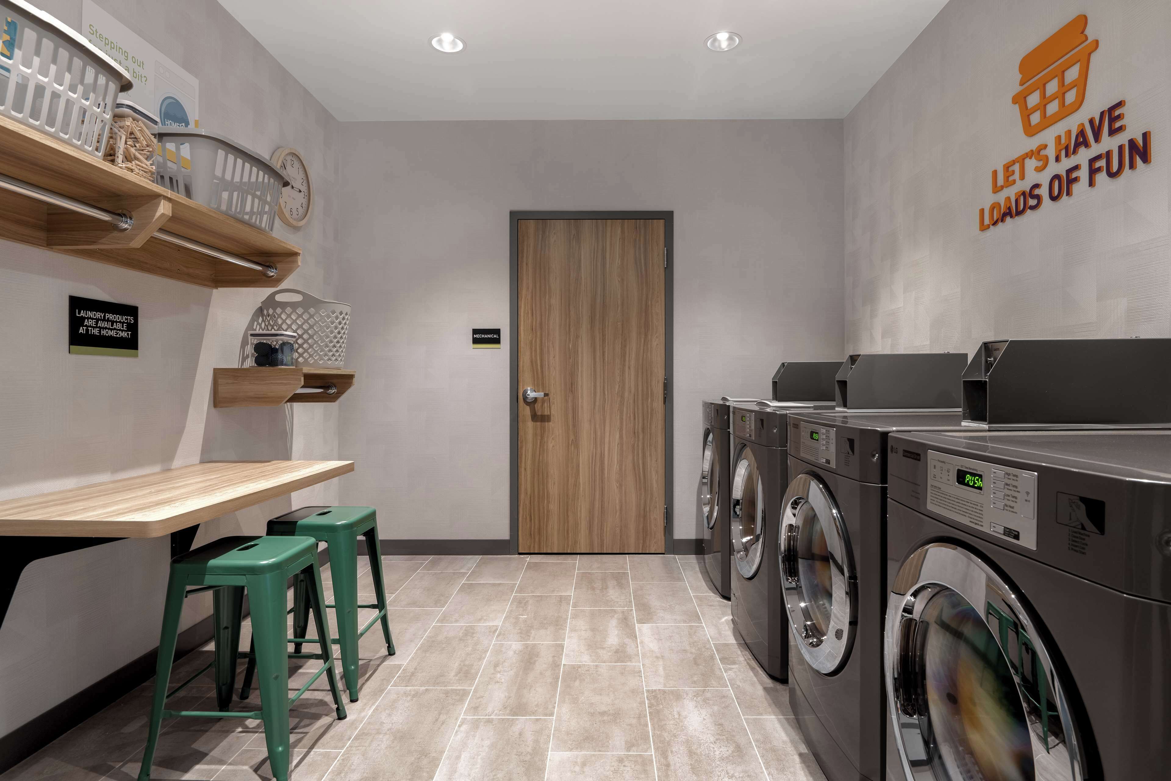 Un lavadero con puerta de madera, dos lavadoras, una mesa con taburetes y estantes con cestas de ropa.
