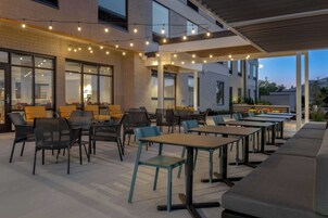 Un patio al aire libre con mesas y sillas, luces colgantes y vistas del edificio al fondo.