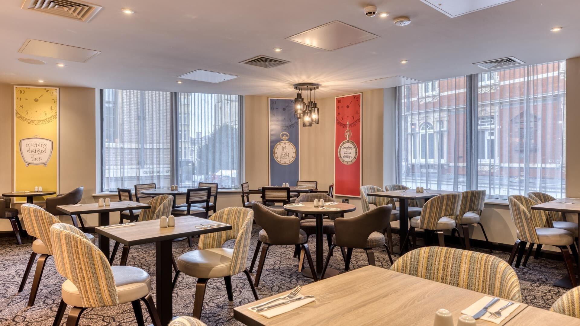 A hotel dining area with multiple tables and chairs, large windows, and decorative wall art.