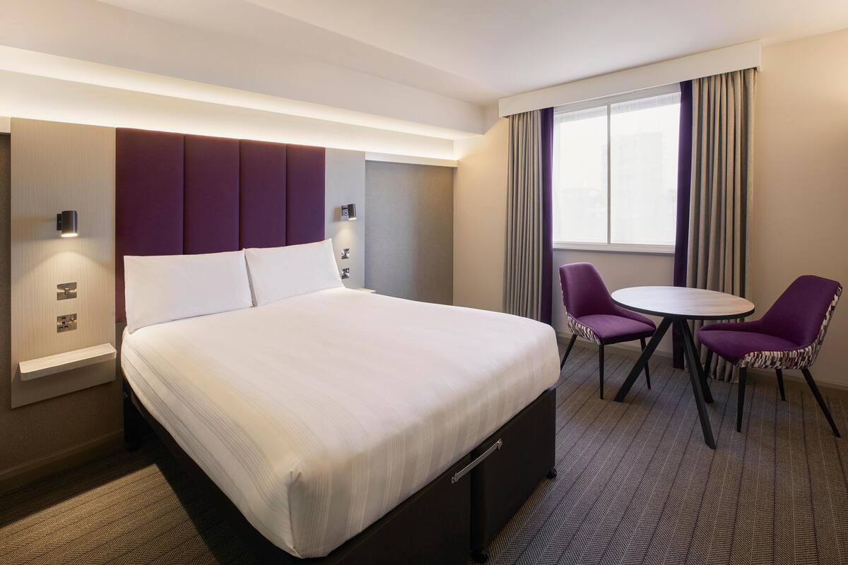 Une chambre d’hôtel avec un grand lit, deux fauteuils violets, une petite table et une fenêtre avec des rideaux.