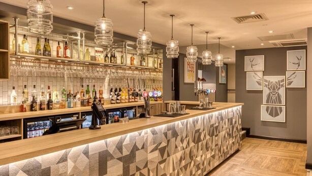 Un espace bar avec un comptoir, des étagères remplies de bouteilles et de verres, et un barman.