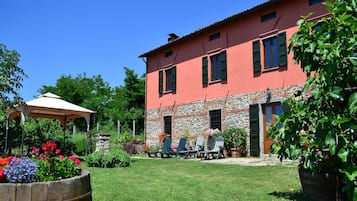 Il s’agit d’un bâtiment à deux étages, doté d’une façade en pierre et d’une couleur rouge. On y trouve un patio avec une table et des chaises, ainsi qu’un jardinier en bois rempli de fleurs.