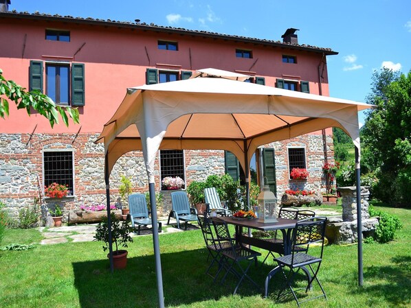 Un patio con mesa y sillas bajo una pérgola, un muro de piedra y un edificio rosa con ventanas y contraventanas.