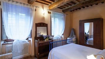 Une chambre avec un plafond en bois, un lit recouvert d’une courtepointe blanche, une commode, un miroir et une fenêtre ornée de rideaux légers.
