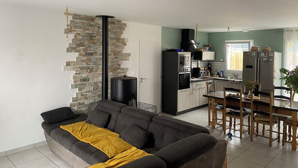 A modern living room with a grey sofa and a yellow throw blanket, a kitchen with a black stove and white cabinets, and a dining area with wooden chairs and a table. - Homestay for rent.  House in a very quiet subdivision. (Les Essarts)