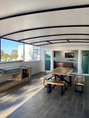 Un espacio moderno para comer al aire libre con mesa y bancos de madera, una parrilla empotrada y un ventanal con vistas a la ciudad.