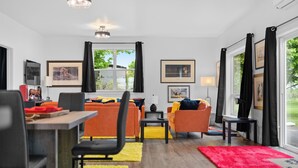 Een moderne woonkamer met een bank, stoelen en een eettafel. Er hangen afbeeldingen in lijsten aan de muren en een groot raam met gordijnen.