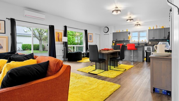 Een modern appartement met woonkamer, eethoek en keuken. Er is een groot raam met gordijnen, een bank met kussens en een eettafel met stoelen.