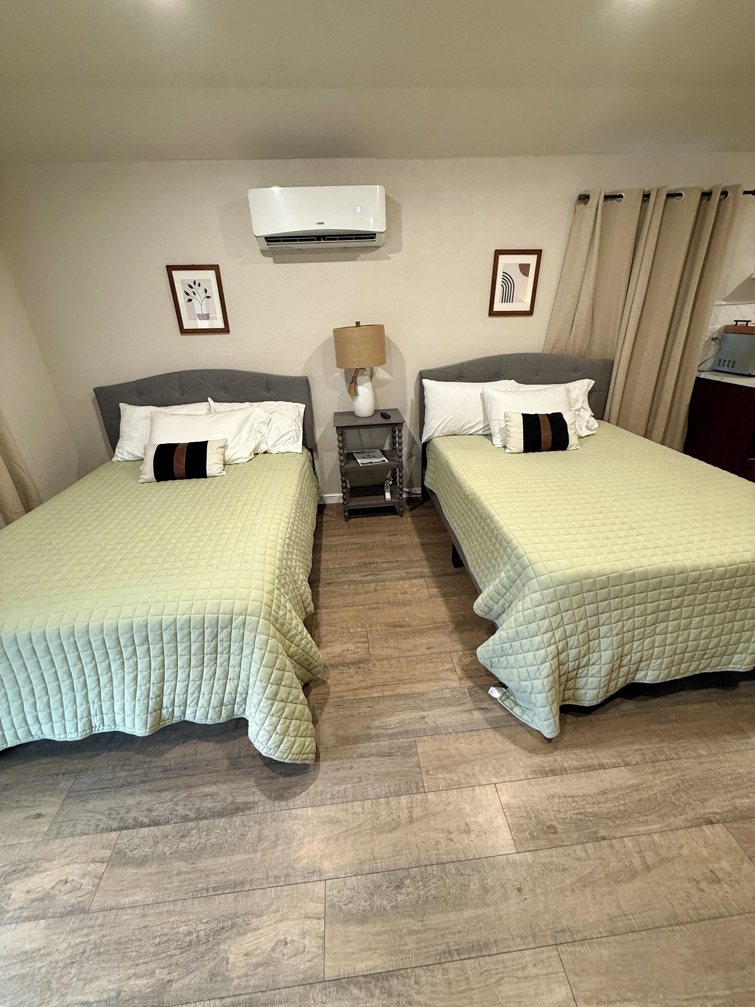 Hotellihuone, jossa on kaksi sänkyä, yöpöytä lampun kanssa, ilmastointilaite ja seinällä oleva kehystetty kuva.