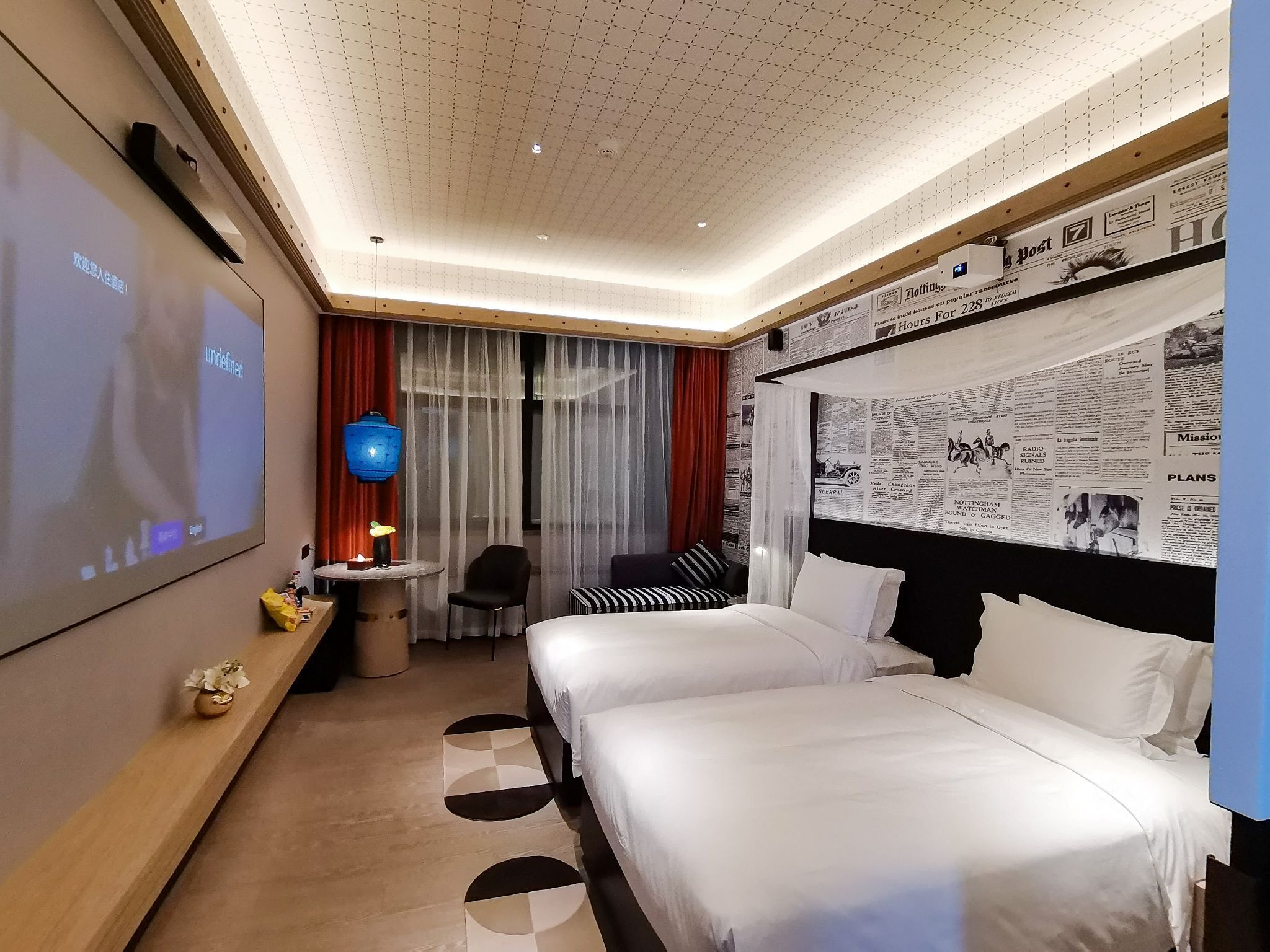 Ein Hotelzimmer mit einem großen Bett, einem Schreibtisch mit Lampe, einem Fernseher und einer Wand mit Zeitungsausschnitten.