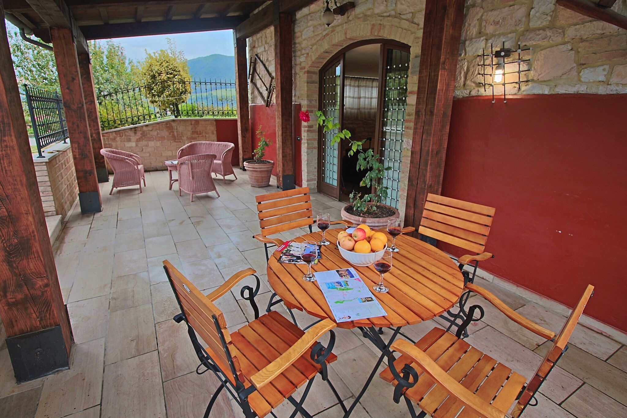 A covered patio with wooden furniture, a table with fruit and wine glasses, and a view of the outdoors.