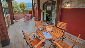 A covered patio with wooden furniture, a table with fruit and wine glasses, and a view of the outdoors.