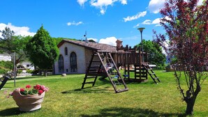A playground with a wooden swing set and climbing structure, a large flower pot, and a building with a tiled roof in the background.