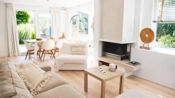 Een moderne woonkamer met een bank, salontafel en een open haard. Er is een eethoek met stoelen en een tafel, en een raam met uitzicht op groen.