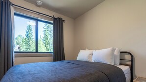A bedroom with a large window, a bed with a dark bedspread, white pillows, and a wooden nightstand. - Newly Built 2 Bedrooms. Sleeps 4 Adults. AC (Fernie)