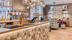 A bar area with multiple beer taps, a counter with a cash register, and a seating area with a plaid chair and ottoman. - Premier Inn Dublin Cc North Docklands (Dublin)