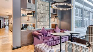 A modern lounge area with a plush purple sofa, a round table, and a bookshelf filled with books. - Premier Inn Dublin Cc North Docklands (Dublin)