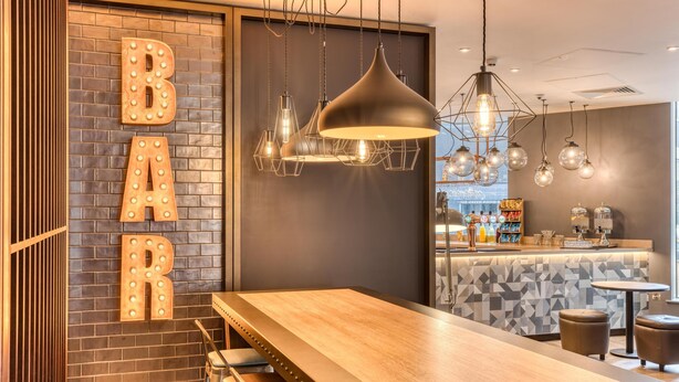 A bar area with a wooden bar counter, hanging pendant lights, and illuminated letters spelling 'BAR' on the wall. - Premier Inn Dublin Cc North Docklands (Dublin)
