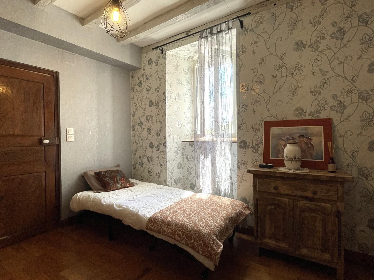 Um quarto pequeno com uma cama de solteiro, um guarda-roupa de madeira, uma janela com cortinas florais e um quadro pendurado na parede.